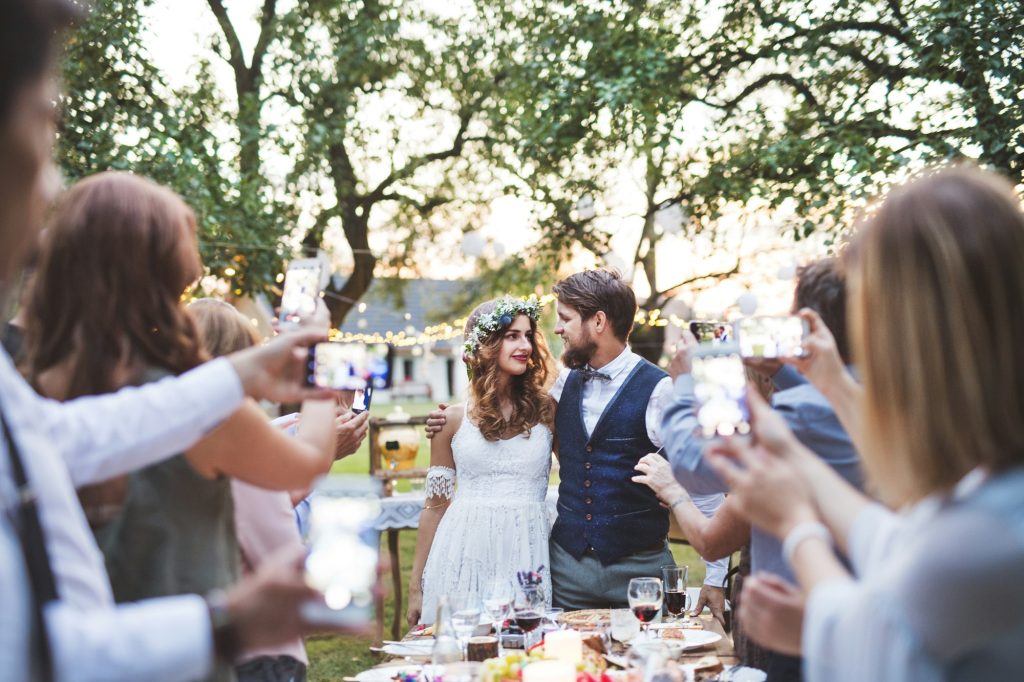 Guests with smartphones taking photo of bride and groom at wedding reception outside.